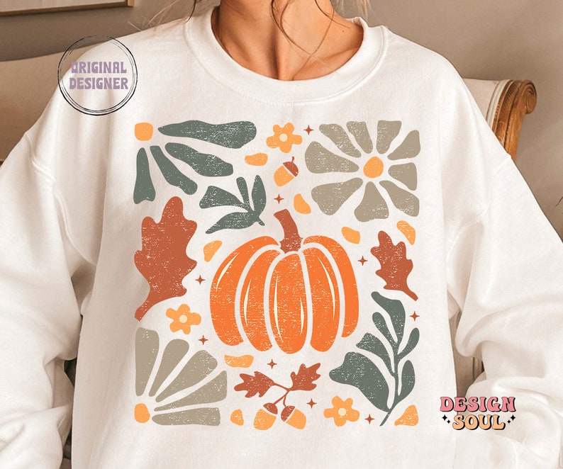 May include: White sweatshirt featuring a graphic design of a pumpkin and autumn foliage in shades of orange, brown, and green. The design is in a square shape and has a vintage aesthetic.