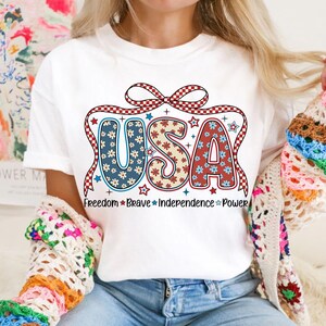 May include: White t-shirt with a red, white, and blue USA graphic. The letters are decorated with a floral pattern and a red and white checkered ribbon. The text "Freedom Brave Independence Power" is printed below the graphic.