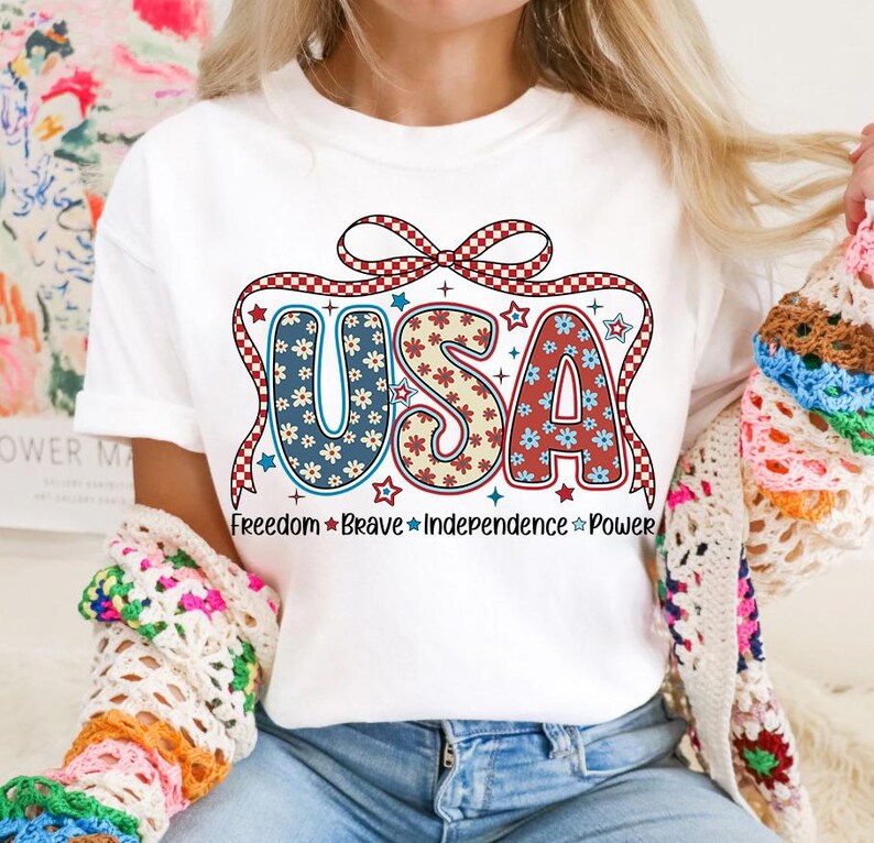 May include: White t-shirt with a red, white, and blue USA graphic. The letters are decorated with a floral pattern and a red and white checkered ribbon. The text "Freedom Brave Independence Power" is printed below the graphic.