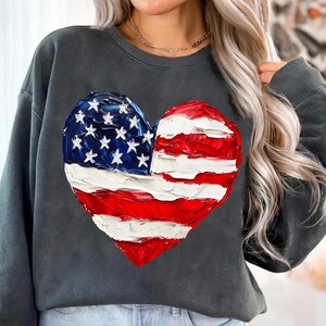 Pu&ograve; includere: Una felpa grigia scura con un design a forma di cuore della bandiera americana dipinto. La bandiera &egrave; rossa, bianca e blu.