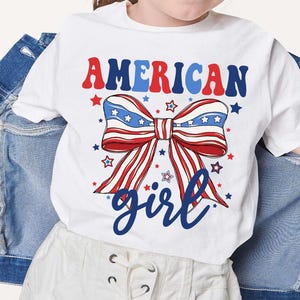 May include: White t-shirt with a red, white, and blue bow with stars and the text "AMERICAN girl" in blue.