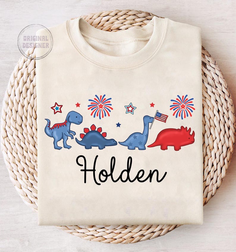 May include: Cream-colored sweatshirt featuring a row of cartoon dinosaurs in blue and red, with fireworks and stars. The name "Holden" is written in black script below the dinosaurs. The upper left corner has the text "ORIGINAL DESIGNER".