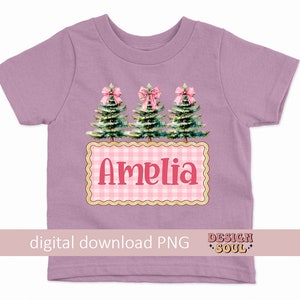以下が含まれることがあります： ピンクのボウと緑のクリスマスツリー3本がデザインされたパープルのTシャツ。ピンクと白のギンガムチェックの四角形にピンク色の「アメリア」という名前が書かれています。