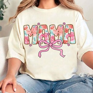 May include: A white t-shirt with a floral design that spells out "MAMA" with a pink bow underneath.