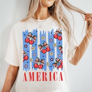 May include: White t-shirt with a blue and red graphic print of cherries and stars. The graphic has the word "AMERICA" printed below it.