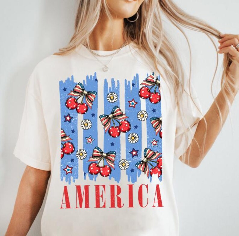 May include: White t-shirt with a blue and red graphic print of cherries and stars. The graphic has the word "AMERICA" printed below it.
