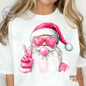 May include: A white t-shirt featuring a pink and white illustration of Santa Claus wearing pink heart-shaped sunglasses and blowing a pink bubble. Santa is giving the peace sign with his right hand.
