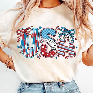 May include: A white t-shirt with a red, white, and blue "USA" graphic. The letters are decorated with different patterns, including stars, stripes, and polka dots.