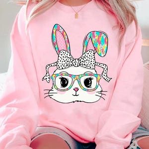 May include: Pink sweatshirt with a white bunny wearing glasses and a polka dot bow. The bunny's ears are patterned with colorful geometric shapes.