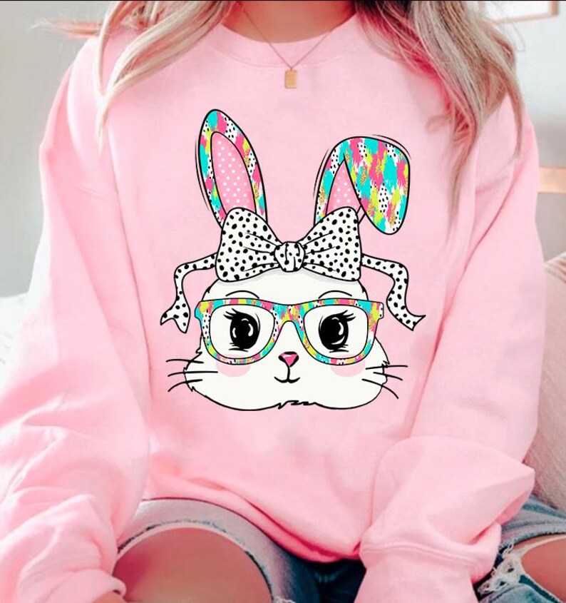 May include: Pink sweatshirt with a white bunny wearing glasses and a polka dot bow. The bunny's ears are patterned with colorful geometric shapes.
