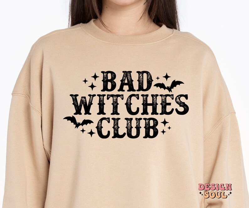 May include: A beige sweatshirt with the text "BAD WITCHES CLUB" in a distressed black font. The design includes bat and star graphics. The sweatshirt has a crew neck and long sleeves.