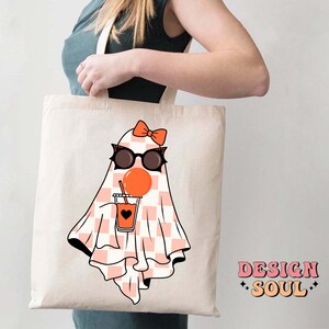 May include: A white tote bag with a pink and white checkered ghost wearing sunglasses and a pink bow, holding a cup of orange juice. The text "DESIGN SOUL" is printed on the bag.