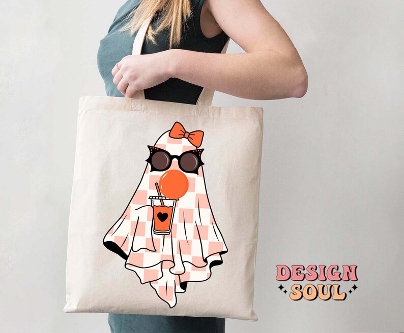 May include: A white tote bag with a pink and white checkered ghost wearing sunglasses and a pink bow, holding a cup of orange juice. The text "DESIGN SOUL" is printed on the bag.