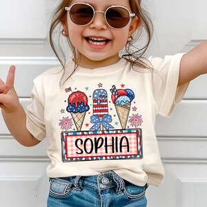 May include: A cream-colored t-shirt featuring a colorful graphic of ice cream cones and popsicles in red, white, and blue. The name "SOPHIA" is displayed in a red and black plaid frame. The shirt is worn by a young child.