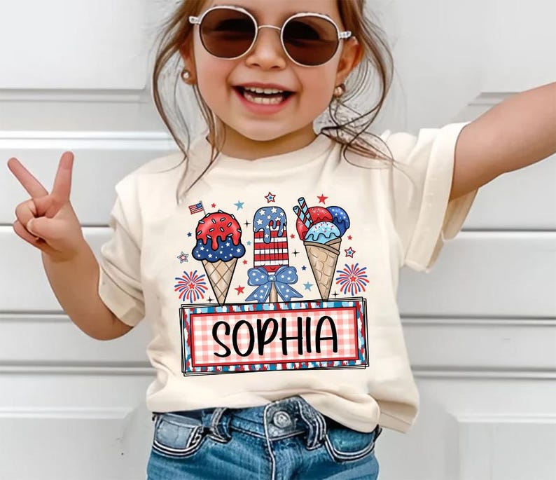 May include: A cream-colored t-shirt featuring a colorful graphic of ice cream cones and popsicles in red, white, and blue. The name "SOPHIA" is displayed in a red and black plaid frame. The shirt is worn by a young child.