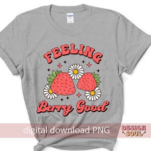 May include: A gray t-shirt with the text "Feeling Berry Good" in a retro font. The text is surrounded by a design of two red strawberries with green leaves and white flowers.