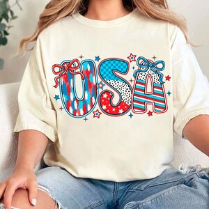 May include: A white t-shirt with a red, white, and blue USA graphic. The letters are decorated with stars, stripes, and polka dots. The graphic includes two red bows.