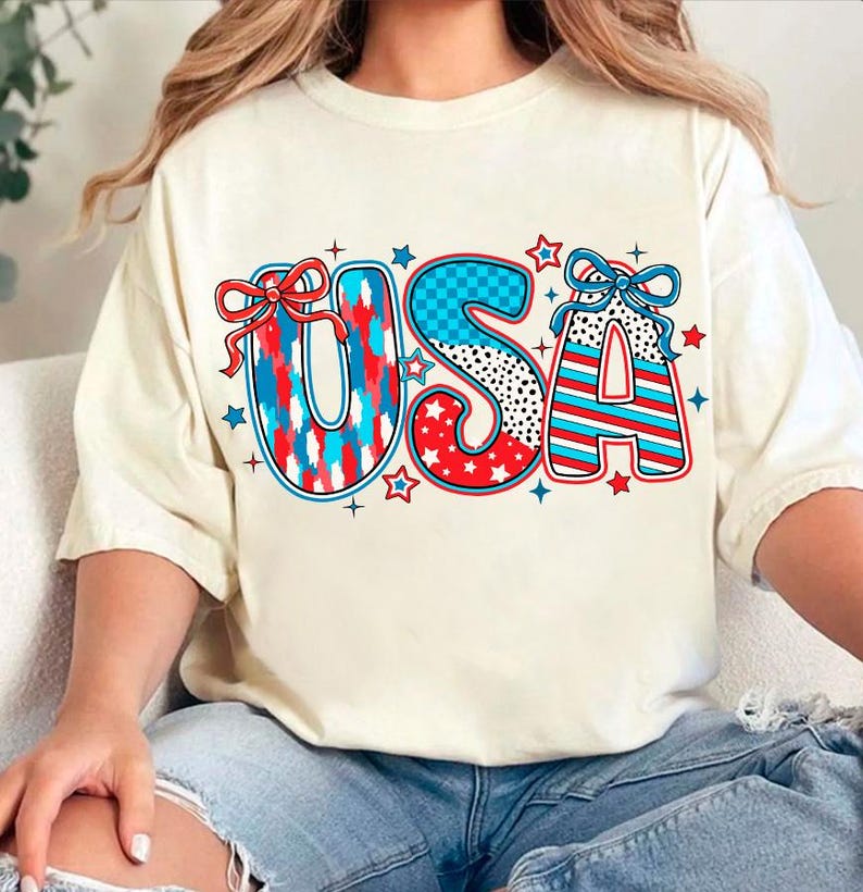 May include: A white t-shirt with a red, white, and blue USA graphic. The letters are decorated with stars, stripes, and polka dots. The graphic includes two red bows.