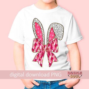 May include: A white t-shirt featuring a pink and gold bunny ear design. The bunny ears are adorned with pink paw prints and black polka dots. The bunny ears are tied together with a pink and gold bow.