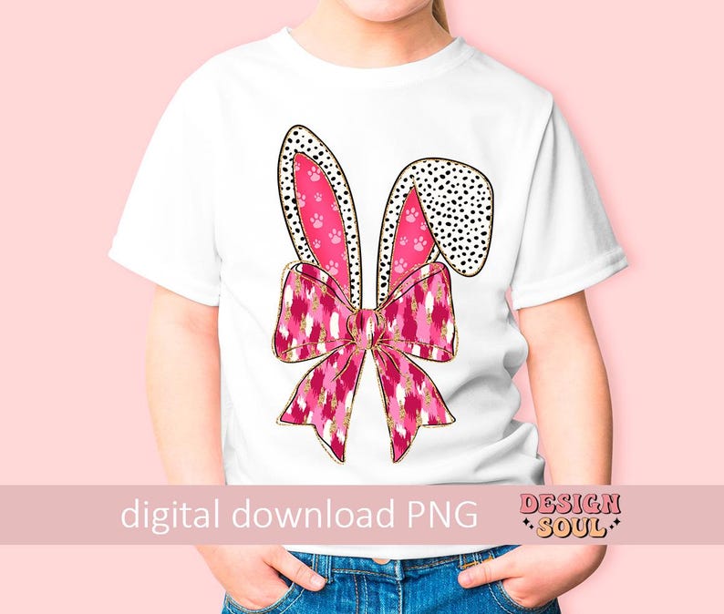 May include: A white t-shirt featuring a pink and gold bunny ear design. The bunny ears are adorned with pink paw prints and black polka dots. The bunny ears are tied together with a pink and gold bow.