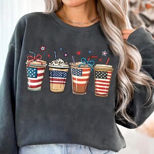 May include: A dark gray sweatshirt with a graphic of four iced coffee cups decorated with the American flag. Each cup has a straw and a red, white, and blue ribbon.