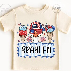 May include: Cream-colored t-shirt featuring a cartoon goose wearing a patriotic hat and sunglasses, with ice cream cones and fireworks. The name "BRAYLEN" is displayed in a blue and white frame. A fun, festive design for kids.