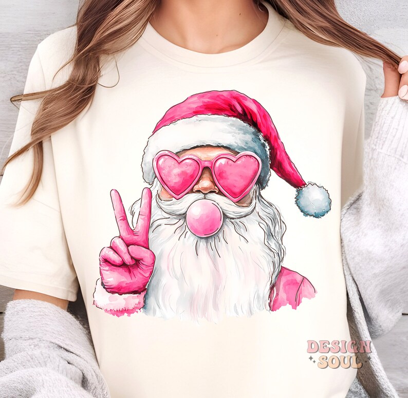 May include: A white t-shirt featuring a pink and white illustration of Santa Claus wearing heart-shaped sunglasses and blowing a pink bubble. Santa is giving the peace sign with his right hand.