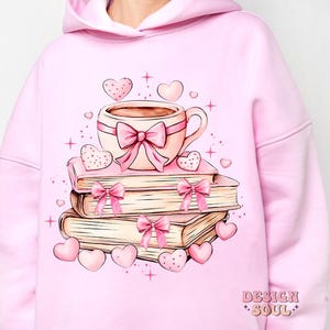 K&ouml;nnte beinhalten: Rosafarbener Hoodie mit einem Motiv aus einer Tasse Kaffee, einem Stapel B&uuml;chern und Herzen. Der Text "Design Soul" ist in der rechten unteren Ecke des Hoodies gedruckt.