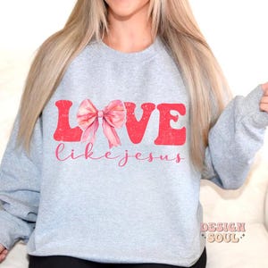 May include: A grey sweatshirt with the words "LOVE like jesus" printed in pink and red. A pink bow is in the middle of the word "LOVE".