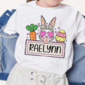 May include: White t-shirt with a pink bunny rabbit wearing sunglasses and holding a lollipop. The bunny is surrounded by pink flowers, a green carrot, and a yellow and white Easter egg. The text "Raelynn" is on a pink and white checkered banner.