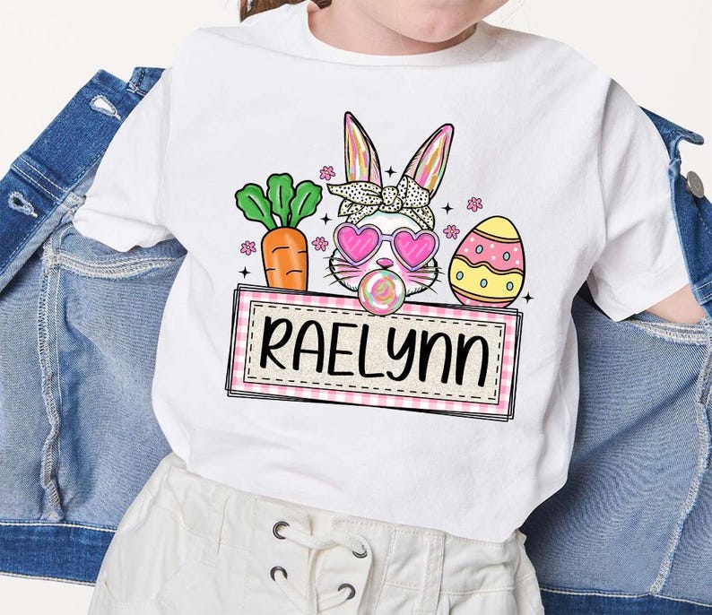 May include: White t-shirt with a pink bunny rabbit wearing sunglasses and holding a lollipop. The bunny is surrounded by pink flowers, a green carrot, and a yellow and white Easter egg. The text "Raelynn" is on a pink and white checkered banner.