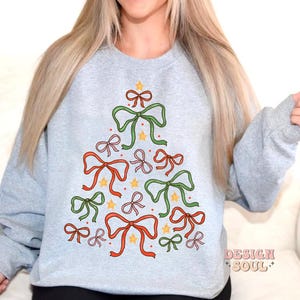 May include: A grey sweatshirt with a Christmas tree design made of colourful bows and stars. The tree is centred on the front of the sweatshirt.