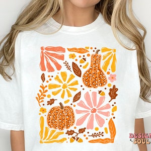 May include: White t-shirt with a graphic print of orange, yellow, pink, and brown pumpkins and flowers. The text "DESIGN SOUL" is printed in the bottom right corner.