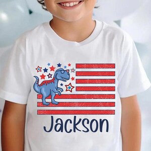 May include: White t-shirt with a red, white, and blue American flag design featuring a blue dinosaur wearing sunglasses. The name "Jackson" is printed below the flag.