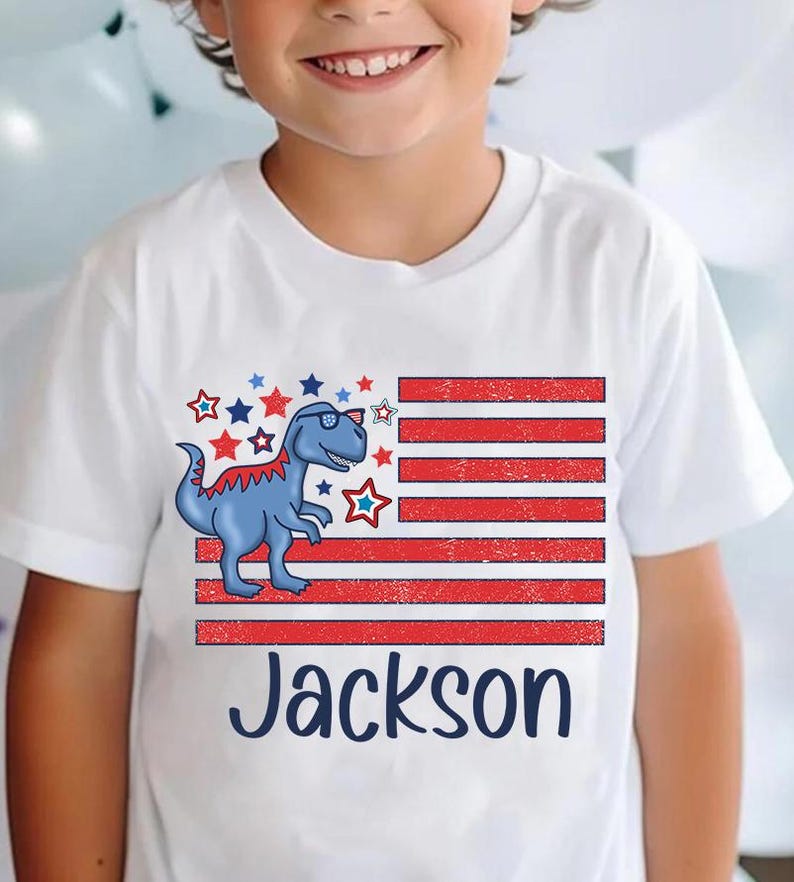 May include: White t-shirt with a red, white, and blue American flag design featuring a blue dinosaur wearing sunglasses. The name "Jackson" is printed below the flag.