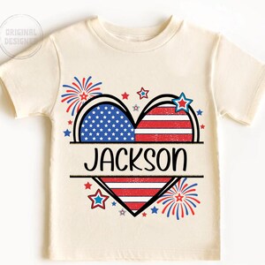 May include: Cream-colored t-shirt featuring a heart design split into two halves, one with blue stars on a white background and the other with red and white stripes. The name "JACKSON" is printed in black. Red, white, and blue stars and fireworks complete the patriotic theme.