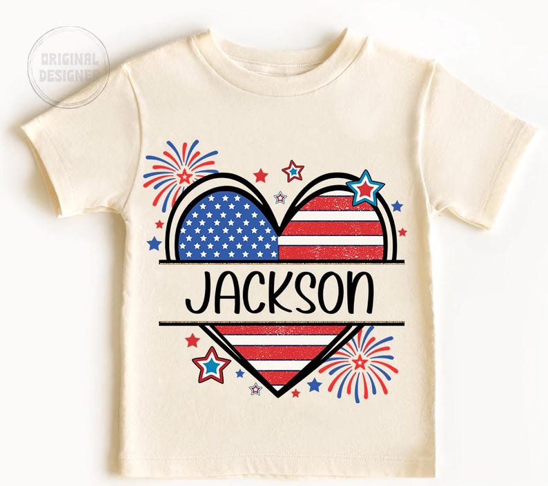 May include: Cream-colored t-shirt featuring a heart design split into two halves, one with blue stars on a white background and the other with red and white stripes. The name "JACKSON" is printed in black. Red, white, and blue stars and fireworks complete the patriotic theme.