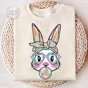 May include: A white sweatshirt featuring a colourful bunny rabbit wearing heart-shaped sunglasses and a polka dot bow. The bunny is holding a colourful lollipop.