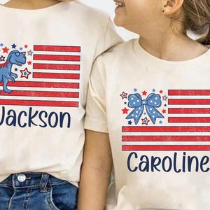 May include: Two white t-shirts with a red, white, and blue American flag design. One shirt features a blue dinosaur with the name "Jackson" printed below. The other shirt features a blue bow with the name "Caroline" printed below.