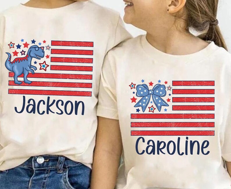 May include: Two white t-shirts with a red, white, and blue American flag design. One shirt features a blue dinosaur with the name "Jackson" printed below. The other shirt features a blue bow with the name "Caroline" printed below.
