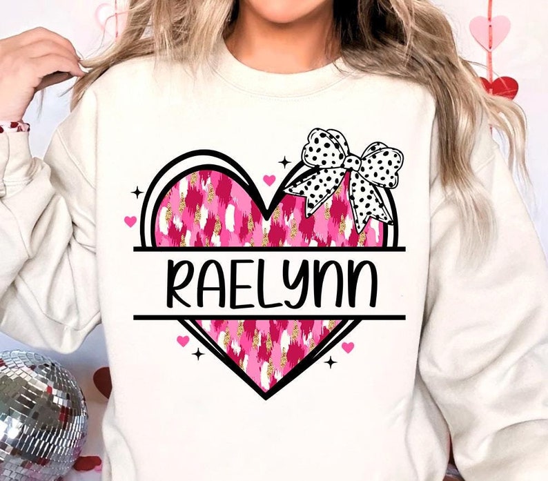 May include: A white sweatshirt with a pink and gold heart design and a black and white bow. The heart has the name "Raelynn" written across it.