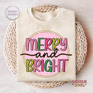 May include: A cream-colored sweatshirt with the words "MERRY and BRIGHT" in pink, green, and black lettering. The text is inside a pink polka-dotted circle with a gold glitter outline. The words "Original Designer" and "Design Soul" are also visible.