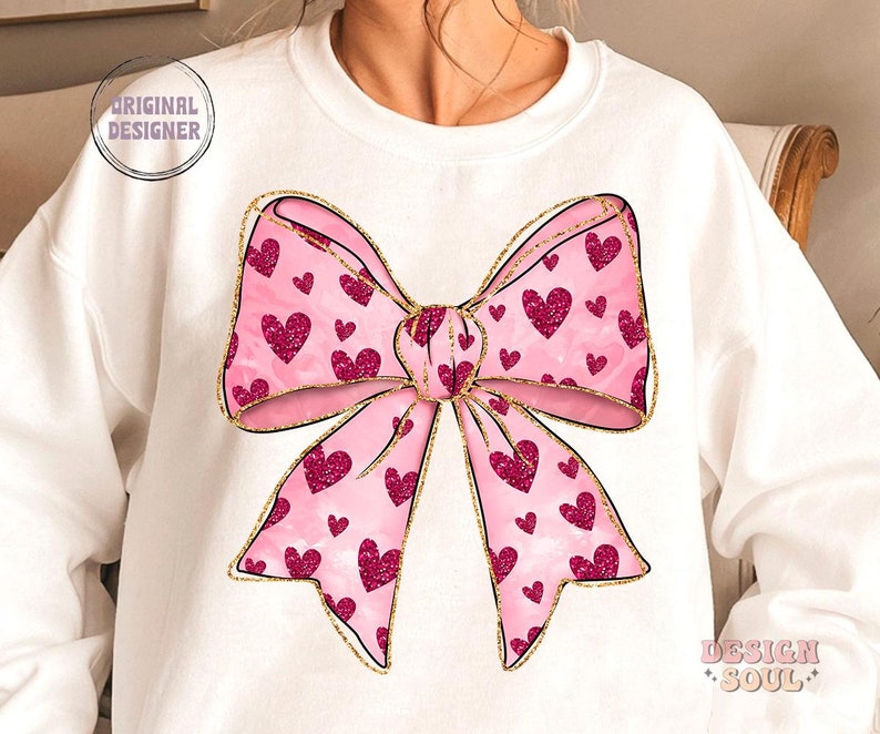 May include: A white sweatshirt with a pink and gold bow with a heart pattern. The bow is centered on the front of the sweatshirt.