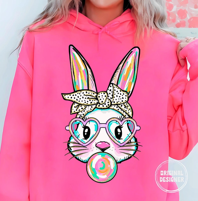 May include: A pink hoodie featuring a graphic of a white bunny wearing heart-shaped sunglasses and a polka dot headband. The bunny is blowing a bubblegum bubble.