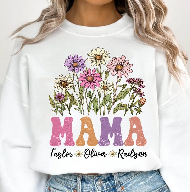 May include: White sweatshirt featuring a floral design with daisies in shades of pink, purple, and yellow. The word "MAMA" is printed in large, retro-style letters, with the names "Taylor", "Oliver", and "Raelynn" below.