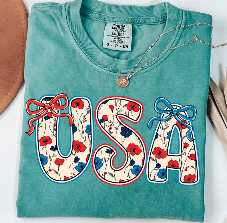 May include: A teal blue t-shirt with a white, red, and blue floral pattern spelling out "USA". The shirt has a distressed look and a bow on each letter.
