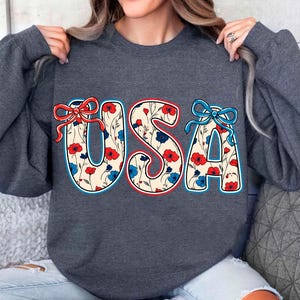 May include: A dark gray sweatshirt with a floral pattern "USA" graphic. The letters are outlined in blue and red, and the flowers are red, blue, and white.