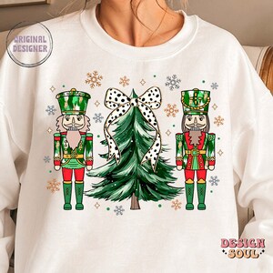 May include: White sweatshirt with a festive Christmas design. It features two nutcrackers in green and red uniforms, a green Christmas tree with a black and white polka dot bow, and snowflakes. The words "ORIGINAL DESIGNER" and "DESIGN SOUL" are also visible.