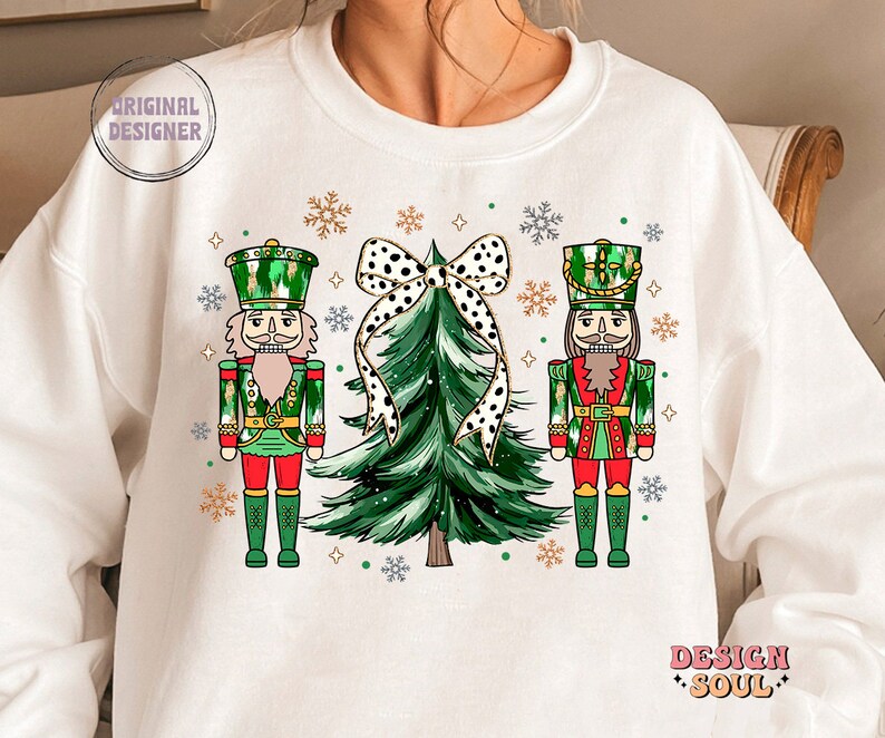 May include: White sweatshirt with a festive Christmas design. It features two nutcrackers in green and red uniforms, a green Christmas tree with a black and white polka dot bow, and snowflakes. The words "ORIGINAL DESIGNER" and "DESIGN SOUL" are also visible.