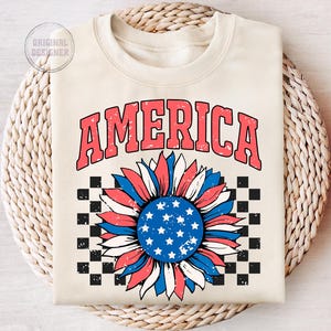 May include: A cream-colored sweatshirt with the word "AMERICA" in red and blue letters. A red, white, and blue sunflower with a checkered background is below the word.
